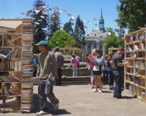 An outdoor space with wooden bookshelves around which several people are browsing and selecting books. There is an old colonial building in the distance with a bell tower.