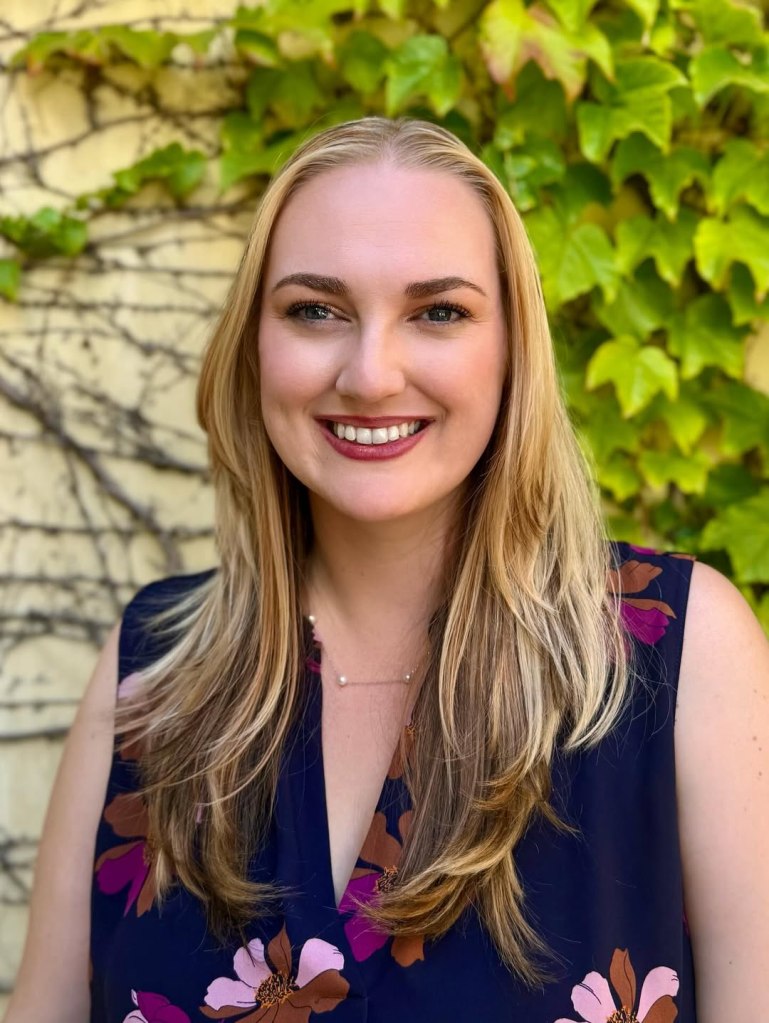 Author Rebecca Robinson's portrait. She has blond hair and dark eyes, and wears a blue dress with pink and brown flowers on it. In the background is a wall covered in ivy.