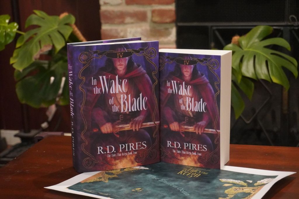 Hardcover and paperback copies of the fourth book in The Tides That Reign epic fantasy series, In the Wake of the Blade. They are posed standing vertically on a wooden table atop a canvas map of the world in the series.